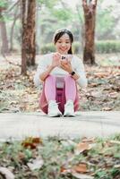 sentado mujer en rosado polainas y blanco zapatillas usos su teléfono inteligente en un parque camino, con auriculares en y arboles en el antecedentes. foto