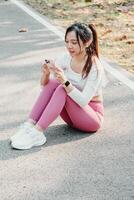 sentado mujer en rosado polainas y blanco zapatillas usos su teléfono inteligente en un parque camino, con auriculares en y arboles en el antecedentes. foto