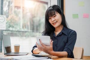 sonriente mujer de negocios con tirantes utilizando calculadora a su escritorio con un ordenador portátil y café en un brillante espacio de trabajo foto