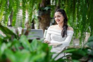 Woman smiles as she works on her laptop, seated comfortably in a garden surrounded by hanging ferns and greenery. photo