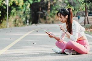 joven mujer en rutina de ejercicio engranaje disfruta un romper, utilizando su teléfono inteligente en un soleado parque la carretera. foto