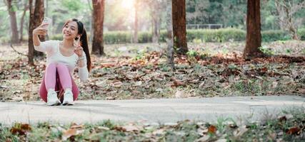 Active young woman in sportswear happily captures a selfie on a tree-lined park path, displaying a playful peace sign. photo