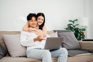 Young Asian couple enjoys a cozy moment on their sofa, interacting with a laptop, in a light-filled, plant-adorned living space. photo