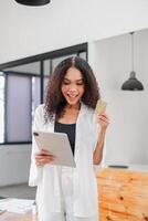 Smiling businesswoman in a white blazer holding a credit card and tablet, likely managing online transactions in her office. photo