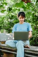 Smiling young woman sitting on a wooden bench, working on her laptop in a lush green garden setting. photo
