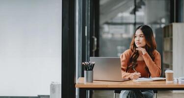 Creative professional sits thoughtfully at her work desk with a laptop, pondering over new ideas in a bright office. photo