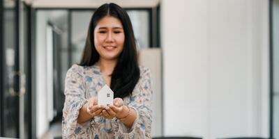 Smiling woman in a floral dress holds a small wooden house model. photo