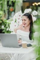 Laughing heartily, a young woman enjoys a call on her laptop in a cafe surrounded by lush greenery, exemplifying a joyous work-life balance. photo