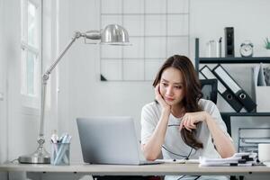 Young businesswoman appears reflective while seated at her bright and orderly home office, with a laptop and documents on her desk. photo