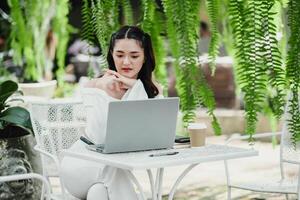 Young woman is absorbed in her work on a laptop at a white table in an outdoor cafe surrounded by greenery. photo