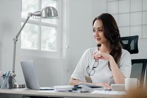 Professional woman with glasses in hand, working in a well-lit home office with a laptop and desk lamp. photo