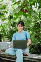 Happy young woman working on a laptop, seated outdoors on a wooden bench surrounded by green foliage. photo