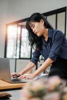 Serious Asian businesswoman concentrating on her work on a laptop at a busy office desk, with paperwork. photo