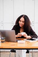 Cheerful businesswoman enjoying a cup of coffee during a work break, casually using her laptop in a bright office. photo