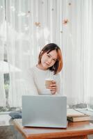 joven mujer por casualidad participación un café taza, tomando un momento descanso desde trabajar, sentado en frente de su ordenador portátil en un brillante, aireado habitación. foto