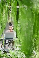 Cheerful woman working on her laptop, framed by vibrant hanging ferns at a garden cafe. photo