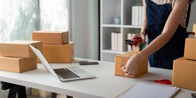 Woman use scotch tape to attach parcel boxes to prepare goods for the process of packaging at home, shipping, online sale internet marketing ecommerce concept startup business idea photo