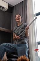 House cleaning with fun. Happy young asian housewife singing song during cleanup, using mop as guitar, enjoying domestic work. Young woman dancing and cleaning in living room photo