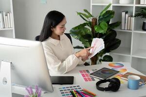 asiático mujer Lanza libre gráfico diseñador trabajando con color muestra de tela muestras y computadora a escritorio en hogar oficina, joven dama elegir color gama para nuevo diseño proyecto foto