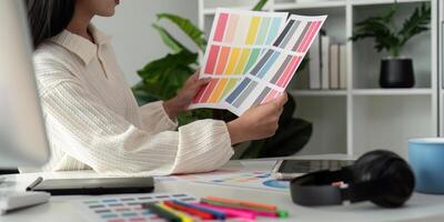 Asian woman freelance graphic designer working with color swatch samples and computer at desk in home office, young lady choosing color gamma for new design project photo