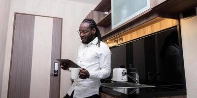 African American man using digital tablet at the kitchen reading social media internet, typing text or shopping online. Freelancer remotely working online at home photo