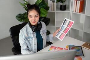 Asian woman freelance graphic designer working with color swatch samples and computer at desk in home office, young lady choosing color gamma for new design project photo