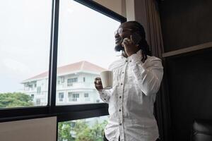 African American man using have call by phone while drink coffee by the window in the living room photo