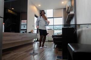 gracioso africano americano es limpieza casa haciendo tareas del hogar limpieza piso con fregona y escuchando a música en inalámbrico auriculares, africano americano hombre es bailando y canto foto