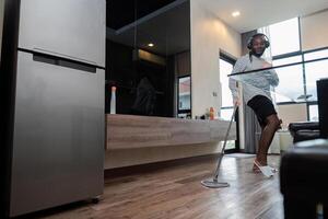 Young African American having fun with mop and music at home, showing dance moves and singing in living room. African American man mopping wooden floors and doing spring cleaning, housework photo