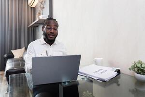 africano americano hombre trabajando con ordenador portátil computadora remoto mientras sentado a vaso mesa en vivo habitación. negro chico hacer Lanza libre trabajo a hogar oficina foto