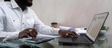 African American man working with laptop computer and using calculator, making financial audit, reviewing bills tax and accounting in living room. Black guy do freelance work at home office photo