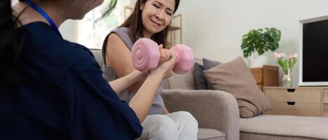 Senior elderly Asia woman with medical caregiver or physical therapist helping patient holding dumbbell in physical therapy session at home. Healthy old people concept photo