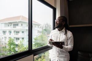 Happy young African business man using online app on tablet for job communication, looking out the window photo