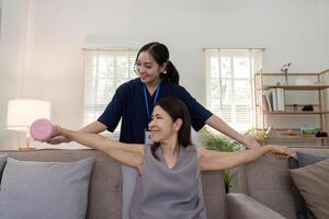 antiguo mujer formación con fisioterapeuta utilizando pesas a hogar. terapeuta asiático ayudando mayor mujer con ejercicio en enfermero hogar. mayor paciente utilizando pesas con extendido brazos foto