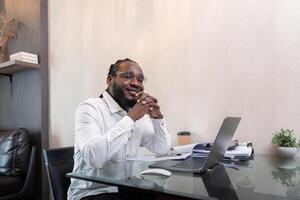 Smiling African American man sit at desk in home working on laptop, happy African American male look at camera posing, busy using modern computer gadget at home photo