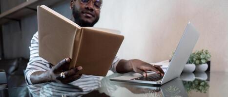 joven africano hombre utilizando ordenador portátil a hogar, negro masculino mirando a leer libro relajante en ocio con trabajo sentar en vaso mesa en vivo habitación foto