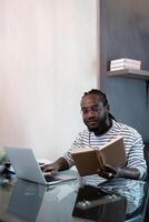 joven africano hombre utilizando ordenador portátil a hogar, negro masculino mirando a leer libro relajante en ocio con trabajo sentar en vaso mesa en vivo habitación foto