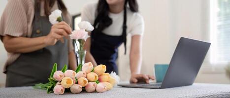 mayor madre y adulto hija contento en el mesa mientras organizar flores en un florero juntos utilizando ordenador portátil computadora. estilo de vida concepto. contento hora juntos foto