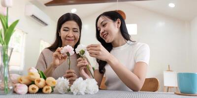 mayor madre y adulto hija contento en el mesa mientras organizar flores en un florero juntos. tecnología y estilo de vida concepto. contento hora juntos foto