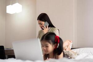 Asian woman in casual outfit is talking on the mobile phone and working on laptop at home with her daughter photo