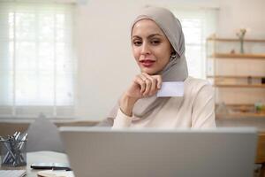 Young happy muslim woman in hijab at home using laptop shopping online with credit card while sitting on desk photo