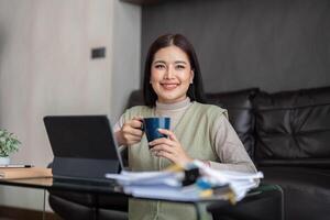 contento joven asiático Lanza libre negocio mujer sentado en el piso a hogar trabajando en digital tableta mientras Bebiendo café foto