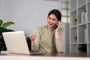 Happy beautiful asian woman relaxing using of laptop computer while sitting on table. Young creative girl working and talk on the phone at home. work at home concept photo