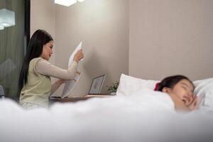 asiático madre trabajando en cama con dormido hija por lado a hogar foto