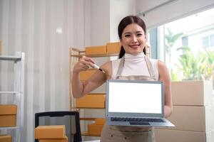 joven negocio mujer asiático trabajando en línea comercio electrónico compras a su tienda. joven mujer vender preparar paquete o empaquetar caja de producto para entregar a cliente. en línea de venta y Bosquejo utilizando ordenador portátil foto