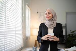 negocio mujer musulmán. confidente mujer de negocios musulmán en hijab en pie con doblada brazos cerca su lugar de trabajo en oficina foto