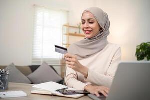 Young happy muslim woman in hijab at home using laptop shopping online with credit card while sitting on desk photo