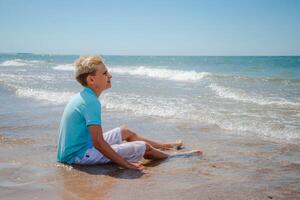 Handsome teenager boy of European appearance with blond hair in white shorts, and a blue T-shirt sits on a beach in sea water and looking away. Summer vacation concept.Summer travel concept.Copy space. photo