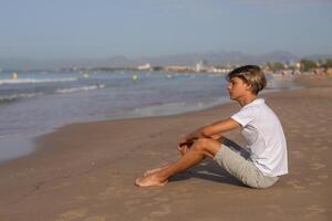 Handsome teenager boy in white t-shirt sitting at the beach and enjoy a summer vacation looking away,Salou city Spain.Family holidays,vacation,trip,travel concept.Side view, copy space. photo