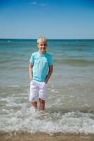 hermoso adolescente chico en blanco pantalones cortos y un azul camiseta soportes en el mar en el agua, y mira a el cámara. verano vacaciones concepto.verano viajes, familia vacaciones, viaje concepto.vertical foto. foto